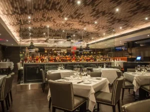 Elegant fine dining restaurant interior with stylish seating, white table settings, and a fully stocked bar area.
