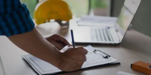 Construction professional reviewing building plans on a clipboard with a laptop and safety helmet nearby.