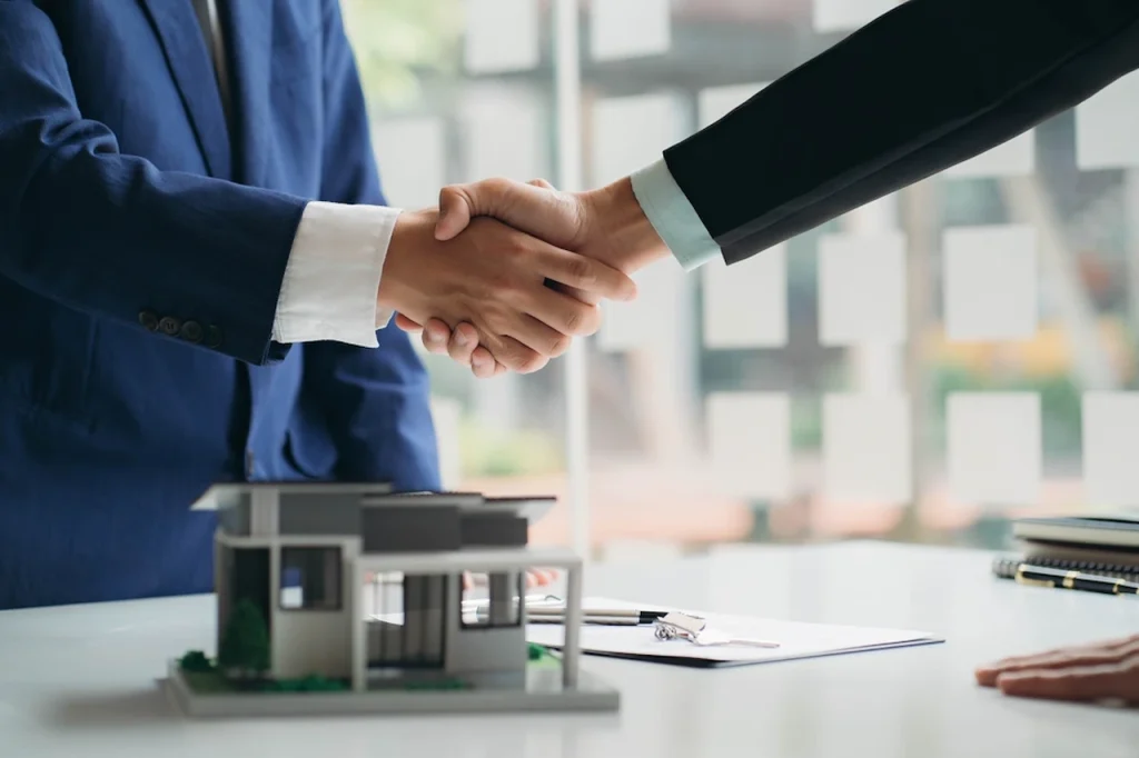 Real estate agents shaking hands over a property deal with a model home and contract documents on the table.
