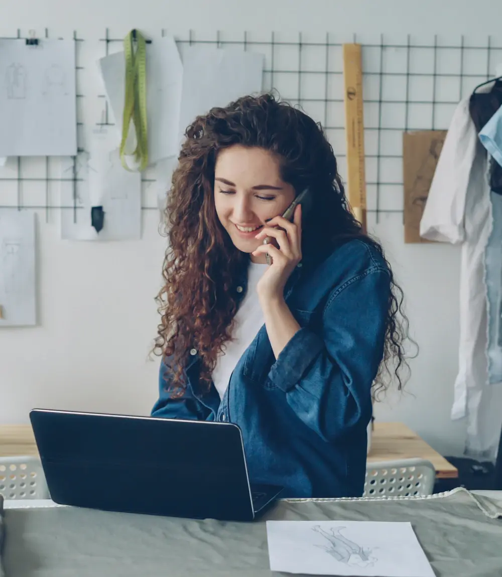 Small business owner answering customer messages on her phone while working on a laptop